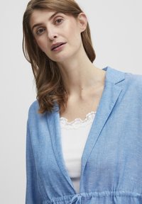 Gaiši zila lina žakete ar brīvu piegriezumu, kas aprīkota ar jostu jostasvietā un šaula apkakli, kas valkāta virs baltas blūzes ar mežģīņu apdari.