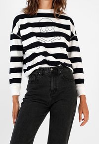 Femme portant un t-shirt à rayures noires et blanches avec un design de crâne, associé à un jean noir taille haute, sur un fond uni.