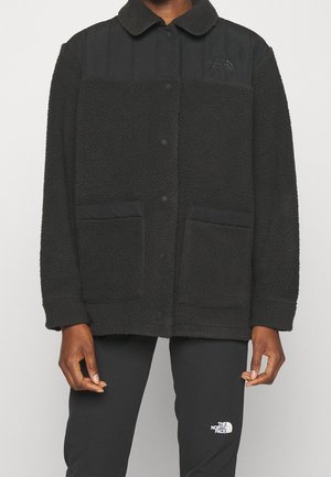 Persona con chaqueta de forro polar negra texturizada con botones a presión y dos bolsillos delanteros, combinada con pantalones negros con el logo de The North Face.