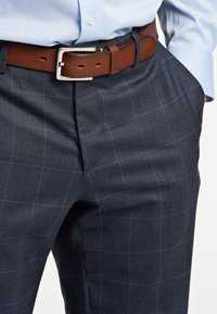 Pantaloni a quadri blu navy in un tessuto testurizzato, con una cintura in pelle marrone dotata di una fibbia in metallo e cuciture a contrasto.