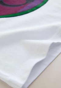 White cotton fabric, smooth texture, showing a folded edge. Features a colorful round design with purple and green accents.
