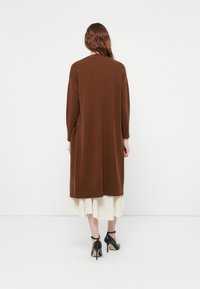 Cardigan lungo marrone con una vestibilità morbida, tasche laterali e orlo a costine, indossato sopra una gonna plissettata bianca. Materiale a maglia testurizzato.