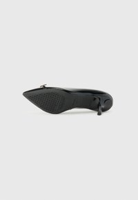Zapato de tacón alto de charol negro con punta afilada y suela de goma perforada. Cuenta con una pequeña correa en el tobillo. Superficie lisa y brillante.