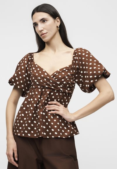 Γυναίκα που φοράει καφέ πουά τοπ με φουσκωτά μανίκια και ραμμένη ακριβώς στο μπούστο, ποζάρει με το ένα χέρι στη μέση μπροστά σε απλό φόντο.