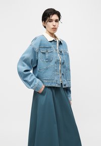 Mladý člověk s krátkými tmavými vlasy, oblečený v světle modré džínové bundě přes béžovou košili s límečkem a tyrkysové kalhoty širokých nohavic, stojí s jednou rukou v kapse.