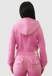 Haut court rose à capuche avec un ourlet côtelé, présentant des détails cousus. Assorti à un pantalon avec des poches arrière décoratives.