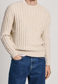 Beige Kabelstrickpullover mit gerippten Bündchen und Saum, über einem weißen Hemd getragen, kombiniert mit blauen Jeans. Glatte Textur und lockere Passform.