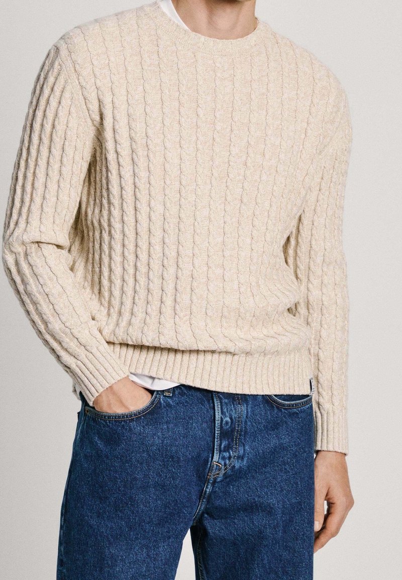 Beige Kabelstrickpullover mit gerippten Bündchen und Saum, über einem weißen Hemd getragen, kombiniert mit blauen Jeans. Glatte Textur und lockere Passform.