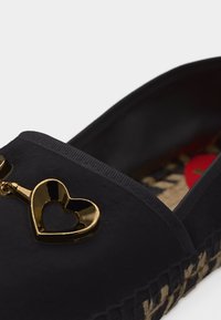 Chaussure noire à enfiler avec semelle tissée et décoration en métal doré en forme de cœur sur le dessus.