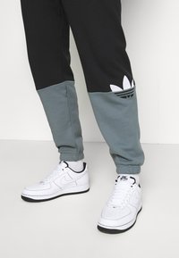 Svart och teal joggers med en avsmalnande design och ett broderat vitt Adidas-logotyp; kombinerade med vita Nike Air Force 1-sneakers.
