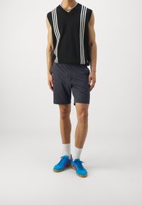 Maglione senza maniche nero con strisce bianche, pantaloni corti grigi, sneakers blu con dettagli rossi e calze bianche. Texture del tessuto liscia nel complesso.