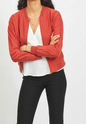 Kobieta ze skrzyżowanymi ramionami, ubrana w rdzawej barwy kurtkę bomberkę, białą bluzkę, czarne spodnie oraz warstwowe złote naszyjniki.