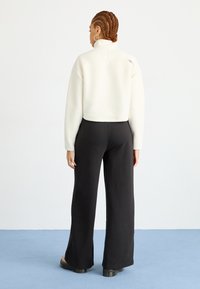 Giacca accorciata in pile bianco con colletto alto, abbinata a pantaloni neri a gamba larga. L'outfit presenta un tessuto strutturato e una vestibilità rilassata.