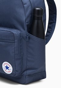 Sac à dos bleu marine en tissu durable, avec une poche latérale contenant une bouteille d'eau noire et un logo Converse circulaire à l'avant.