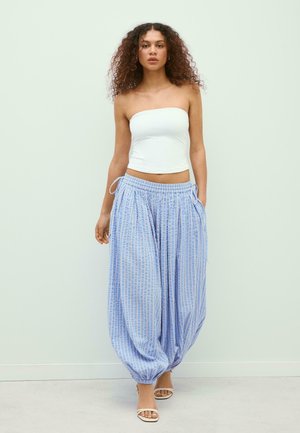 Donna con capelli ricci che indossa un top bianco senza spalline, pantaloni larghi a righe blu e bianche e tacchi bianchi aperti, in piedi contro una parete chiara e semplice.