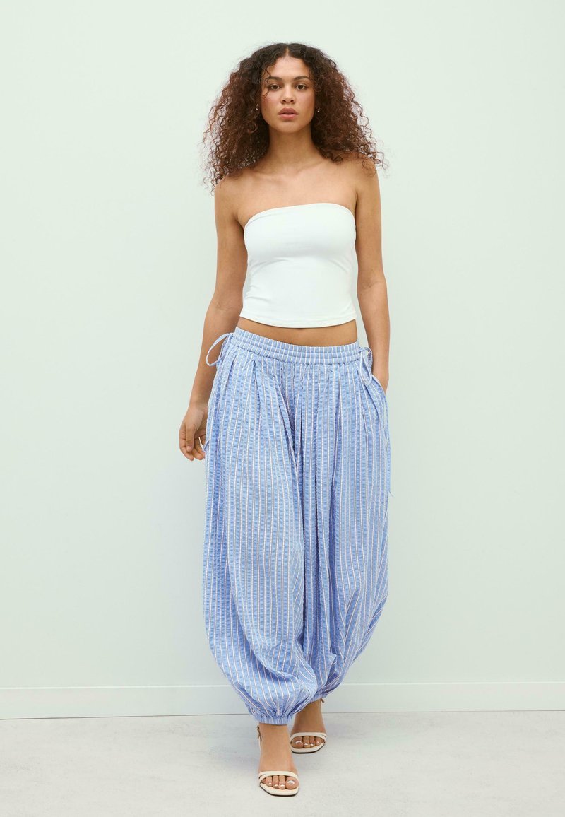 Femme aux cheveux bouclés portant un top blanc sans bretelles, un pantalon ample rayé bleu et blanc, et des talons ouverts blancs, debout contre un mur clair uni.