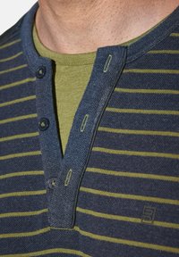 Nahaufnahme einer Person, die ein knopfbares Hemd mit blauen und grünen Streifen über einem olivgrünen Rundhalsunterhemd trägt.