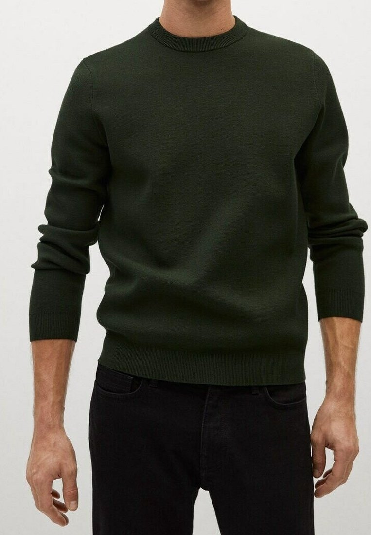 Homme portant un pull col rond vert foncé avec des poignets et un ourlet côtelés, associé à un pantalon noir sur un fond uni.