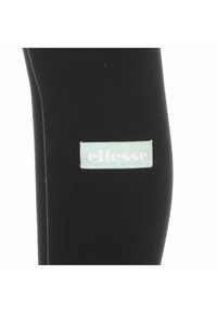 Sorte leggings laget av elastisk stoff med en lys blå rektangulær etikett med ordet "ellesse" i hvitt. Glatt tekstur, tettsittende design.