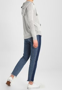 Felpa grigia chiara con cappuccio e maniche lunghe, abbinata a jeans blu con una striscia laterale scura e orli sfrangiati, indossata con sneakers bianche.