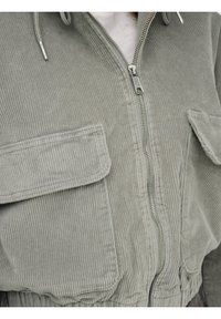 Kurtka ze sztruksu w oliwkowej zieleni z kapturem; posiada przedni zamek błyskawiczny, dwie kieszenie na piersi oraz teksturowaną tkaninę. Regulowany sznurek w talii.