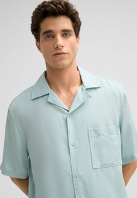 Kurzarm-Hemd mit Kragen in Hellblau, aus glattem Stoff; verfügt über eine Brusttasche und eine Knopfleiste mit passenden Knöpfen.