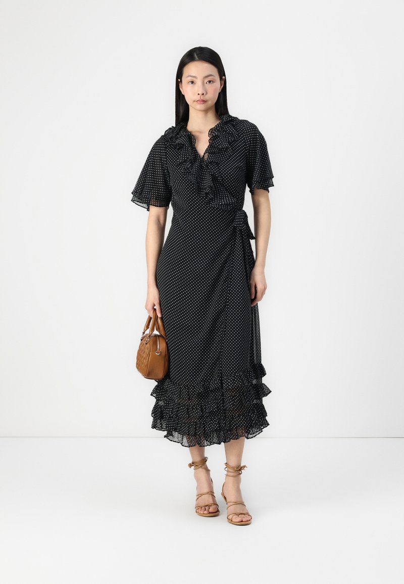 Robe portefeuille noire à pois avec encolure et manches volantes, dotée d'un ourlet à volants en couches. Associée à un sac à main beige et des sandales à lanières.