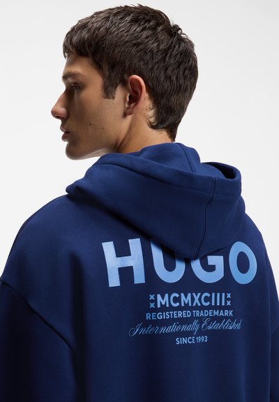Млад мъж в профил носи син суичър с голям светлосин надпис HUGO и допълнителни регистрационни и датни детайли на гърба.