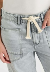 Pantalon en denim gris clair avec une coupe ample, une large ceinture à passants et un cordon de serrage en tissu beige contrastant.