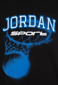 Melnā t-kreklā ir uzraksts "JORDAN" spilgti zilā krāsā ar baltu "sport" zem tā, attēls ar basketbola bumbu un grozu zilā krāsā ar izsmidzinātu efektu.