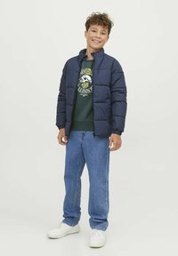Giacca imbottita blu navy con colletto alto e chiusura con zip. Abbinata a una felpa verde, jeans blu e sneakers bianche. Texture liscia su tutta la superficie.