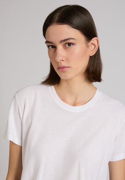 Joven con cabello oscuro y liso, vistiendo una camiseta blanca lisa, mirando ligeramente hacia su izquierda contra un fondo gris claro.