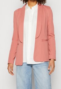 Růžový blazer s klopovým límcem, dvěma předními kapsami, hladkým povrchem, nošený přes bílou košili s knoflíky a světle modrými džíny.