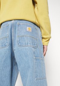 Pantalones vaqueros azul claro con grandes bolsillos traseros y costuras amarillas; presenta un parche con el logo de Carhartt en el bolsillo derecho.