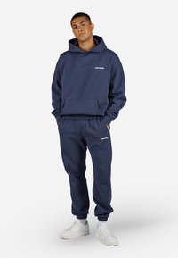Marineblå hettegenser med kengurulomme, elastiske mansjetter og en liten hvit logo. Matchende joggebukser med elastisk midje og ribbestrikkede ankelslutt.