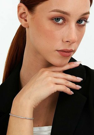 Silberne Armbänder mit kleinen, klaren Steinen, die an einer Hand getragen werden, gegenüber einem schwarzen Blazer. Das Model hat glattes, gerades, rotes Haar und dezentes Make-up.