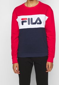 Persoon draagt een rood-wit-blauw FILA-sweatshirt met zwarte broek, staand tegen een effen lichtgrijze achtergrond.