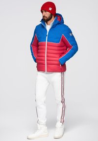 P padded jacka med blå överdel och röd underdel, med vita detaljer. Pairad med vita joggers prydda med röda sidostråk och vita sneakers.