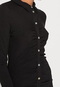 Haut noir à manches longues avec col à revers, fermeture par boutons, et détails froncés sur le côté pour la texture et la forme.