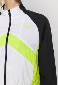 Vit och svart jacka med en djärv neongrön chevrondesign. Har en hög krage och dragkedjestängning, med en liten logotyp på bröstet.