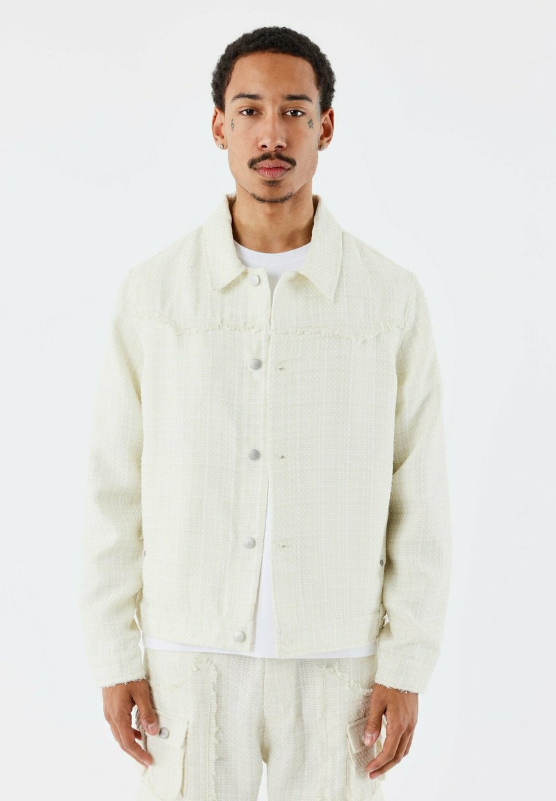 Veste de couleur claire en tissu texturé, avec un fermoir à boutons, un col et un ourlet effiloché. Portée sur une chemise blanche et un pantalon assorti.