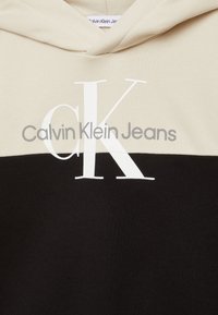 Felpa con cappuccio caratterizzata da una sezione superiore beige e una sezione inferiore nera. Grande logo "CK" bianco e "Calvin Klein Jeans" in grigio. Materiale misto cotone.