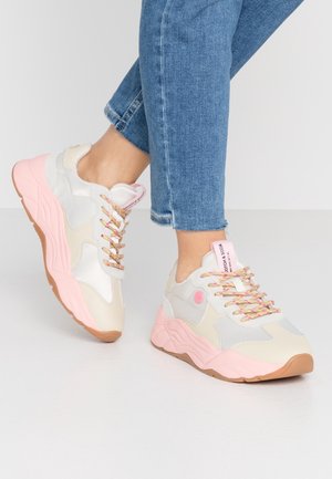 Osoba nosząca jasnobeżowe i różowe masywne sneakersy z kolorowymi sznurówkami oraz niebieskie podwinięte dżinsy na jednolitym tle.