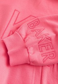 Vaaleanpunainen huppari, jossa on tekstuurillinen brodeeraus kirjaimesta "K" ja sanasta "BAKER" hihassa. Ribbireunat ja pehmeä kangas.