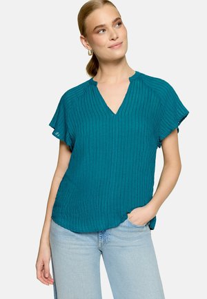 Femme en blouse texturée bleu sarcelle à manches courtes avec col en V et jean bleu clair, la main droite dans la poche, regardant vers le haut.
