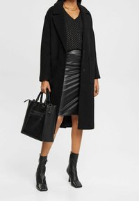 Femme portant un long manteau noir, un haut noir clouté, une jupe en cuir, des bottines et tenant un grand sac à main en cuir noir.