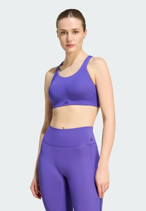 Mujer joven con sujetador deportivo púrpura de Adidas y leggings de talle alto, de pie frente a un fondo gris claro.