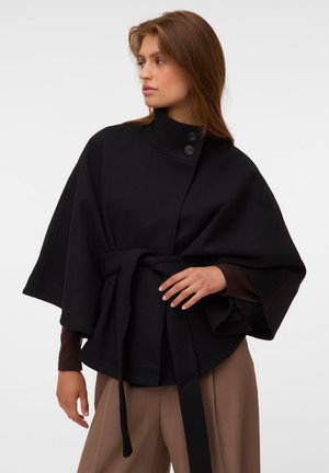 Femme aux cheveux bruns portant une cape noire ceinturée avec deux boutons au col et un pantalon marron, regardant vers sa gauche.