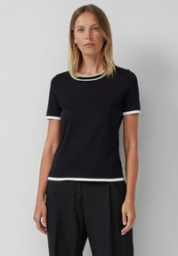 Femme portant un haut noir à manches courtes avec un bord blanc au col, aux manches et à l'ourlet, associé à un pantalon noir, se tenant contre un fond uni.