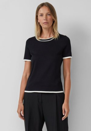 Vrouw in een zwart shirt met korte mouwen en witte afwerking op de kraag, mouwen en zoom, gecombineerd met zwarte broek, staande tegen een effen achtergrond.